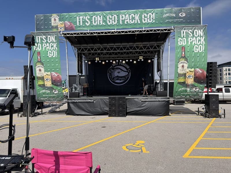 An outdoor show in a parking lot, with PA cabinets and subwoofers stacked in front of the stage under a clear sky.